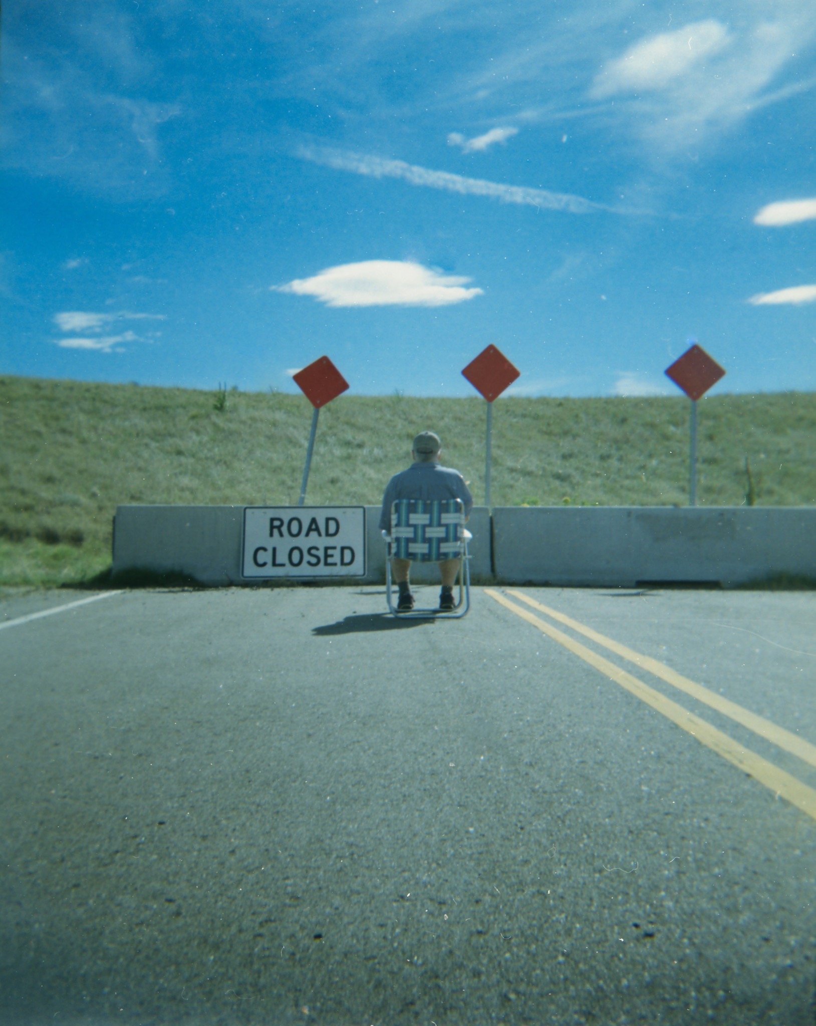 Performance photograph of a seated figure facing a road closed barrier